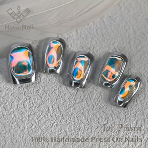 May include: Five iridescent press-on nails with a silver chrome finish. The nails have a holographic effect with shades of blue, green, and orange. The text "Icy Prism 100% Handmade Press On Nails" is visible at the bottom of the image.