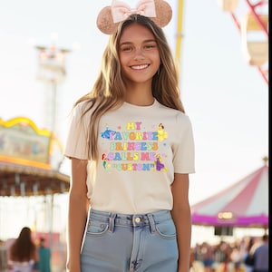 Puede incluir: Una camiseta beige claro con un gráfico colorido que dice "My Favorite Princess Calls Me 'Custon'". El gráfico presenta a varias princesas de Disney en sus colores característicos.