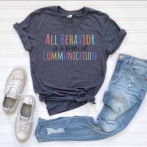 Può includere: Una maglietta grigio scuro con il testo "ALL BEHAVIOR is a form of COMMUNICATION" in colori arcobaleno. La maglietta è abbinata a jeans blu e scarpe da ginnastica argentate.