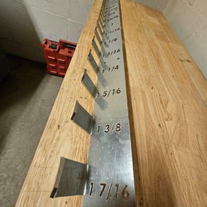 May include: A metal gauge tool with fractional inch measurements. The tool is silver and has cut-out sections for measuring. The measurements are etched into the metal. The tool is on a wooden surface.