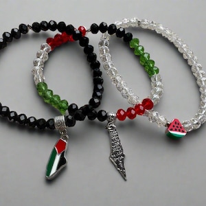May include: Three beaded bracelets with charms. One bracelet has black beads, red and green accents, and a Palestinian flag charm. Another has a silver charm and clear beads with red and green accents. The third has a watermelon charm.