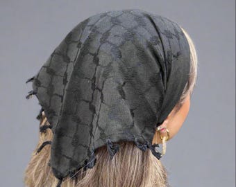 May include: A black keffiyeh headscarf with a geometric pattern, worn over blonde hair. The scarf has fringed edges and is draped over the head and shoulders. The background is a solid gray.