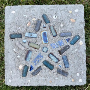 May include: A square concrete stepping stone with a mosaic star design. The mosaic is made of various blue, teal, and white ceramic pieces. Small seashells are embedded in the concrete around the mosaic. The stepping stone is set on a bed of green grass.