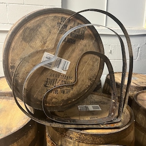 May include: Three metal heart-shaped hoops stacked on top of each other, resting on top of wooden barrels. The barrels are brown and have labels with text that is partially visible. The hoops are made of metal and have a rustic, aged look.