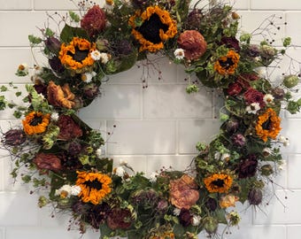 May include: A circular wreath composed of dried sunflowers with vibrant orange petals and dark centers, interspersed with deep red dried roses, green foliage, and delicate white filler flowers, creating a dense, textured botanical arrangement.
