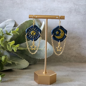 May include: A pair of dangle earrings featuring dark blue wax seal-style charms. One charm displays a gold sun face, the other a gold crescent moon and star. Each charm is accented with a gold-toned metal chain and a small dangling star charm.