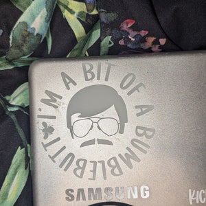 May include: A silver laptop with a white sticker that says "I'm a bit of a bumble but..." with a cartoon image of a man wearing sunglasses and a mustache. The laptop also has the Samsung logo and a black sticker with a symbol.