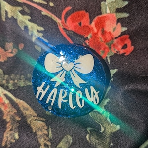 May include: A round, blue glittery object with the name "HARLEY" in white script. A white bow design with a heart in the center is above the name. The object is on a floral patterned background.