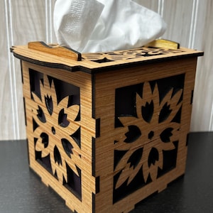 May include: A wooden tissue box holder with a decorative cutout design. The box is made of light brown wood with a dark brown stain. The cutout design features a floral pattern.