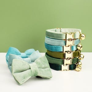 May include: A stack of six velvet cat collars in various shades of green and blue. Each collar has a gold buckle and a small bell. There are also two matching velvet bow ties in a light green color.