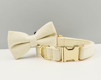 May include: A cream-coloured pet collar featuring a matching fabric bow tie attached to the band. The collar includes a polished gold-tone metal side-release buckle and a matching gold-tone D-ring for attaching a lead, providing a secure closure.