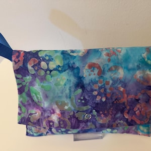 May include: A rectangular fabric clutch featuring a batik-style pattern in shades of blue, purple, teal, and coral. The bag includes a bright blue nylon wrist strap attached to the side and a single silver-toned metal snap closure on the front flap.