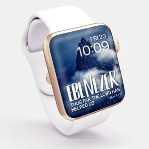 Puede incluir: Reloj inteligente blanco con bisel dorado y correa blanca. La pantalla muestra una escena de montaña con el texto "EBENEZER, THUS FAR THE LORD HAS HELPED US, 1 SAM 7:12" y la fecha y hora.