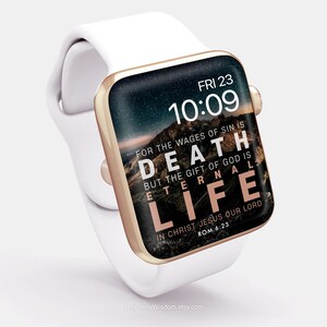 Op de afbeelding: Een witte Apple Watch met een gouden band en een zwart display. Het display toont de tijd, de datum en een Bijbelvers: "Want het loon van de zonde is de dood, maar de gave van God is het eeuwige leven in Christus Jezus, onze Heer. Rom 6:23".