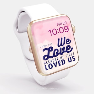 Op de afbeelding: Een witte Apple Watch met een gouden band en een roze en witte aquarel achtergrond. Het horloge gezicht toont de tijd 10:09 en de datum VR 23. De tekst "We Love Because He First Loved Us" wordt weergegeven op het horloge gezicht met de Bijbelverwijzing "1 Johannes 4:19".