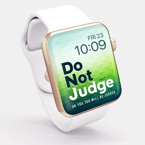 Op de afbeelding: Een witte Apple Watch met een gouden band en een groene en blauwe geometrische achtergrond op het scherm. Het scherm toont de tijd, "FRI 23 10:09", en de tekst "Do Not Judge" met de Bijbeltekst "MATT 7:1" en de zin "OR YOU TOO WILL BE JUDGED".