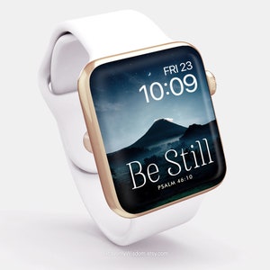 Op de afbeelding: Een witte Apple Watch met een gouden band en een zwart display. Het display toont de tijd, datum en de woorden "Be Still" met de bijbelverwijzing "Psalm 46:10".