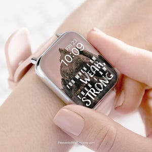 May include: A rose-colored smartwatch with a silver frame displays the time, "FRI 23 10:09", and a mountain graphic. The watch face also features the text "FOR WHEN I AM WEAK, THEN I AM STRONG" and "2 COR 12:10". The watch is on a wrist.
