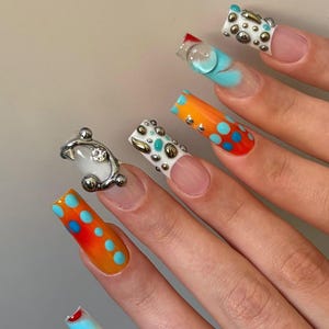 Könnte beinhalten: Lange, quadratisch geformte künstliche Fingernägel mit einer Mischung aus Designs, darunter leuchtend orangefarbene und rote Verläufe mit blauen Punkten, weiße Basen mit silbernen Nieten und transparente 3D-Gel-Tropfen. Ein Nagel trägt einen Metallring.