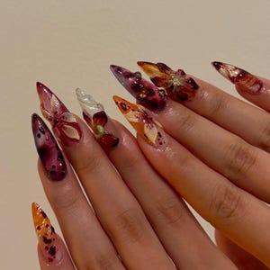 May include: Stiletto-shaped artificial nails featuring a complex 3D design with deep burgundy, orange, and translucent white tones. The nails are decorated with raised floral motifs, dark speckles, and embedded glitter accents across the surface.