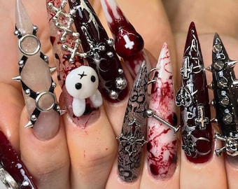 May include: Long stiletto-shaped artificial nails featuring a mix of dark red blood-splatter patterns, black lace designs, and white accents. The nails are decorated with silver metal charms including crosses, skulls, spikes, and a small white bunny figure.