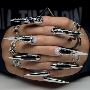 Könnte beinhalten: Lange, spitz zulaufende Nagelverlängerungen in Chromoptik mit schwarzen, flüssig wirkenden Akzenten und Metallnieten. Das Set enthält mehrere silberfarbene Ringe, darunter ein gewundenes Schlangendesign und drahtgewickelte Ringe an den Fingern.