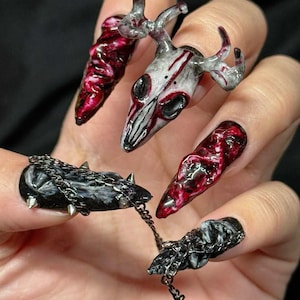 Könnte beinhalten: Ein Set langer, modellierter Kunstnägel mit 3D-Designs. Ein Nagel zeigt einen weißen Hirschschädel mit roten Akzenten. Zwei Nägel haben eine texturierte, glänzende rot-schwarze Marmorierung. Zwei schwarze Nägel sind mit silbernen Nieten und Ketten.