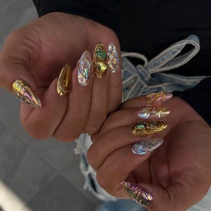 Könnte beinhalten: Lange, mandelförmige künstliche Fingernägel mit einer Mischung aus metallisch goldenen, irisierenden und transparenten Texturen. Einige Nägel enthalten komplizierte Golddrahtwicklungen um grüne und goldene Steine, während andere ein reflektierendes Finish aufweisen.