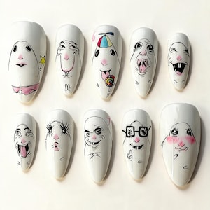 May include: A set of ten white almond-shaped press-on nails, each featuring a unique hand-drawn cartoon character face with distinct expressions, including a lollipop, glasses, and blushing cheeks, rendered in black ink with subtle pink and color accents.