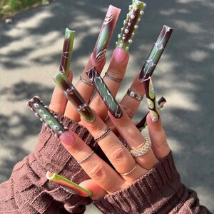 Könnte beinhalten: Nahaufnahme einer Hand mit langen, aufwendig gestalteten Acrylnägeln. Die Nägel weisen eine Vielzahl von Farben auf, darunter Rosa, Grün und Braun, mit dekorativen Elementen wie silbernen Akzenten und Strasssteinen. Die Hand trägt auch mehrere Ringe.