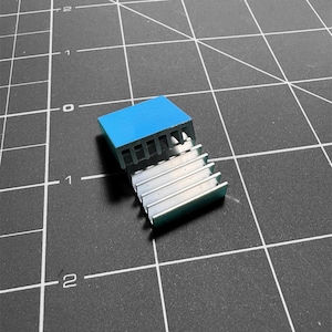 May include: A small, rectangular heat sink with a light blue top and silver fins. The heat sink is made of metal and is designed to dissipate heat from electronic components. It sits on a black cutting mat with white grid lines.