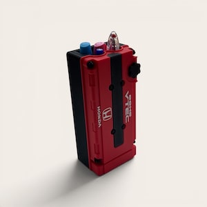 May include: A red and black pen holder shaped like a Honda engine. The pen holder has the Honda logo and the text "DOHC VTEC". It holds several pens and markers in blue, pink, and clear.