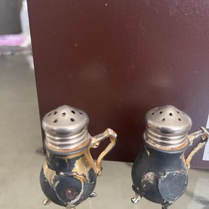 May include: A pair of antique salt and pepper shakers. The shakers have a black base with gold accents and silver tops. Each shaker has a small handle and rests on three small feet. The shakers are displayed on a glass surface.