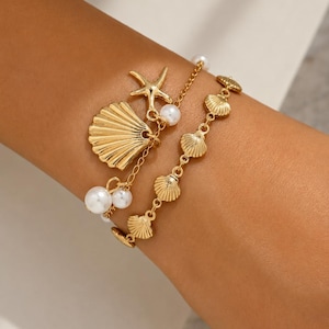 Könnte beinhalten: Goldfarbenes Armband-Set mit Muschel- und Seestern-Anhängern sowie Kunstperlen-Akzenten. Die Armbänder sind geschichtet, eines mit einer Kette aus kleinen Muscheln und das andere mit einem großen Muschel- und Seestern-Anhänger. Das Schmuckstück ist für ein Strand- oder Maritim-Thema konzipiert.