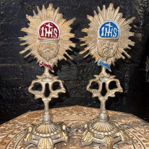 May include: Two metallic gold-toned monstrance-style stands featuring a central sunburst design. Each piece displays a chalice motif with the letters IHS and a cross. One features red enamel accents, while the other features blue enamel accents.
