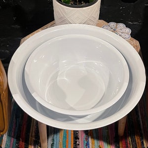 Puede incluir: Dos cuencos de esmalte blanco anidados, uno más grande que el otro, sobre una superficie de madera. Una planta de serpiente en maceta con hojas verdes y amarillas en el fondo. Los cuencos son redondos con un borde ancho.