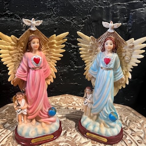 Puede incluir: Dos figuras de ángeles, una rosa y otra azul, cada una con alas doradas, un corazón rojo y una paloma. Cada figura tiene una pequeña figura de niño y un globo. La base dice "Archangel Chamuel".