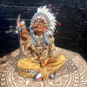 May include: A detailed figurine of a Native American chief in traditional attire, seated with legs crossed. The figure wears a feathered headdress, beaded necklaces, and holds a pipe. The color palette includes tan, blue, red, and white.