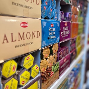 May include: A shelf filled with various incense sticks in colorful boxes. The boxes feature different scents, including Almond, Angel de Mi Guarda, Anti Stress, Camphor, and Cinnamon Patchouli. The boxes are arranged in rows and columns, creating a visually appealing display.