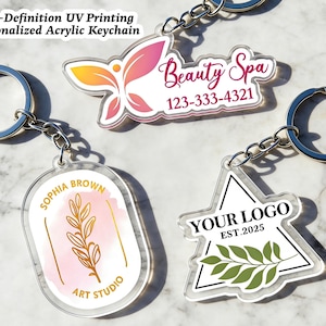 Könnte beinhalten: Transparente Acryl-Schlüsselanhänger mit individuellen Designs. Ein Schlüsselanhänger zeigt ein Blumendesign mit dem Text "SOPHIA BROWN ART STUDIO". Ein anderer hat ein Logo mit dem Text "YOUR LOGO EST.2025". Ein dritter Schlüsselanhänger hat den Text "Beauty Spa 123-333-4321". Jeder Schlüsselanhänger hat einen silbernen Schlüsselring.