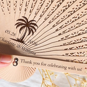 May include: A wooden hand fan with a palm tree design and the text "05.28 Charlotte & William 2026" and "Thank you for celebrating with us!"