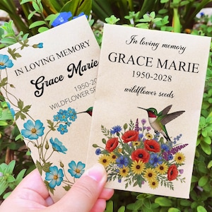 May include: Two memorial seed packets held in hand, featuring floral designs and text. One reads "In Loving Memory Grace Marie 1950-20... Wildflower Seeds" with blue flowers. The other says "In loving memory GRACE MARIE 1950-2028 wildflower seeds" with a hummingbird.