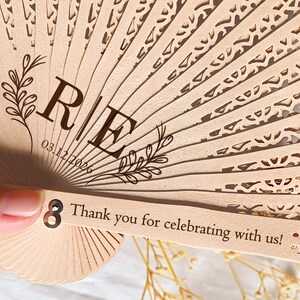 May include: A wooden hand fan with a delicate lace pattern. The fan has a personalized message with the initials "R & E" and the date "03.12.2026". The fan also has the text "Thank you for celebrating with us!" printed on the handle.