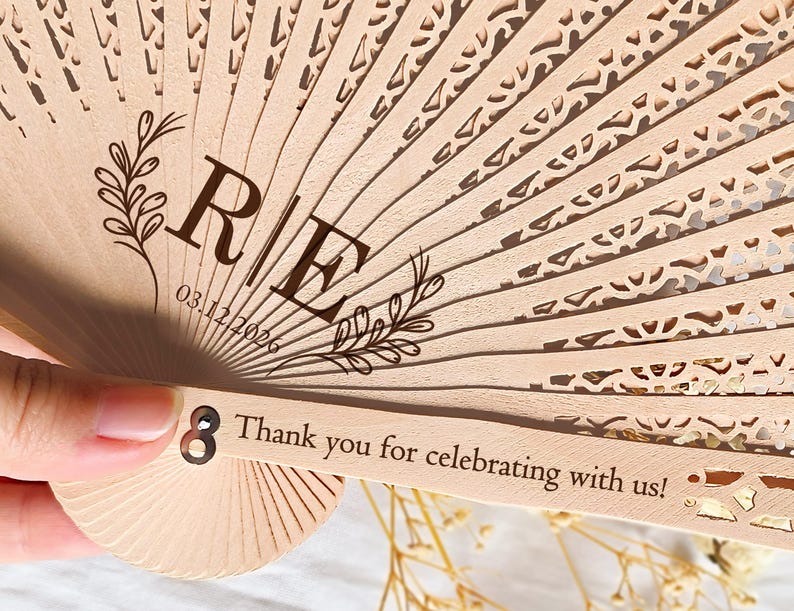 May include: A wooden hand fan with a delicate lace pattern. The fan has a personalized message with the initials "R & E" and the date "03.12.2026". The fan also has the text "Thank you for celebrating with us!" printed on the handle.
