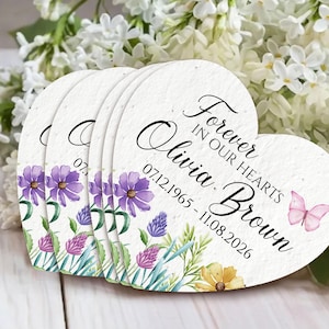 May include: Heart-shaped memorial cards with floral watercolor designs. The cards feature the text "Forever in our hearts" and the name "Olivia Brown" with dates. A pink butterfly and various flowers are also depicted.