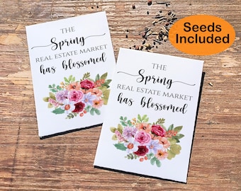May include: Two white cards with floral designs and the text "The Spring Real Estate Market has blossomed" printed on them. The cards are on a wooden surface. Seeds are included.