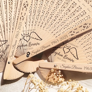 May include: A set of three wooden hand fans with a white lace design. Each fan has a black inscription with the text "Sophia Brown 1963-2025" and a small angel with wings and a halo.