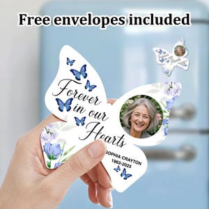 Puede incluir: Una tarjeta conmemorativa blanca con forma de mariposa con detalles florales y acentos de mariposa azul. La tarjeta presenta una foto de una mujer y el texto "Forever in our Hearts". También se incluye el texto "Sophia Crayton 1963-2025".