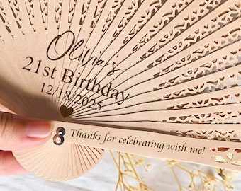 Op de afbeelding: Een lichtbruine houten waaier met ingewikkelde uitgesneden patronen. De waaier is gepersonaliseerd met de naam "Olivia's", "21st Birthday" en de datum "12/18/2025". De tekst "Thanks for celebrating with me!" is ook zichtbaar.