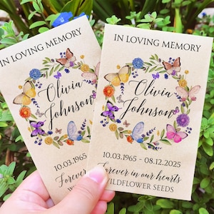 May include: Two brown paper packets with wildflower seeds. Each packet has a floral wreath design with butterflies and the text "In Loving Memory Olivia Johnson" and dates. The text "Forever in our hearts Wildflower Seeds" is printed on the bottom.